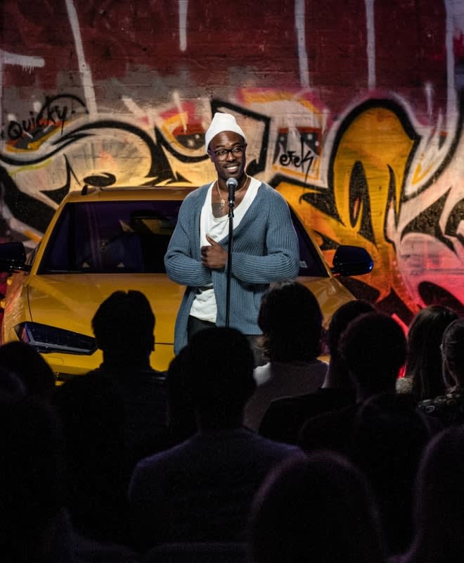 a comedian performing in front of a fancy car and graffiti